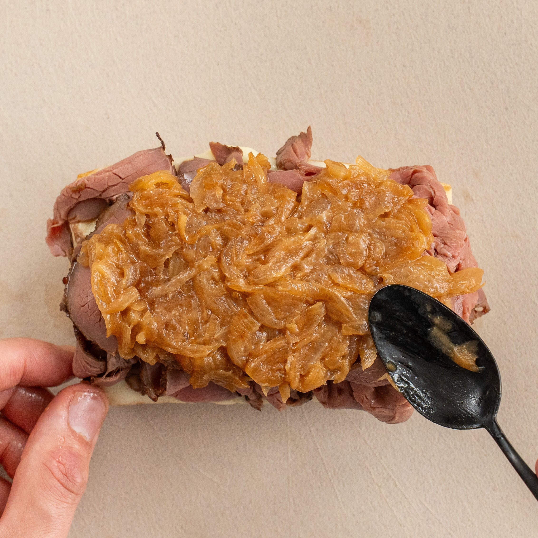 black spoon spreading caramelized onions onto half roast beef sandwich