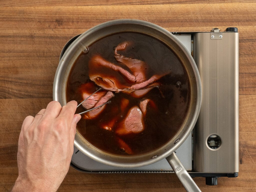 long tweezers stirring sliced roast beef in jus