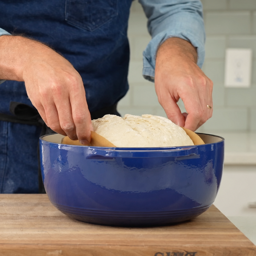 man in blue apron dropping boule into blue dutch oven