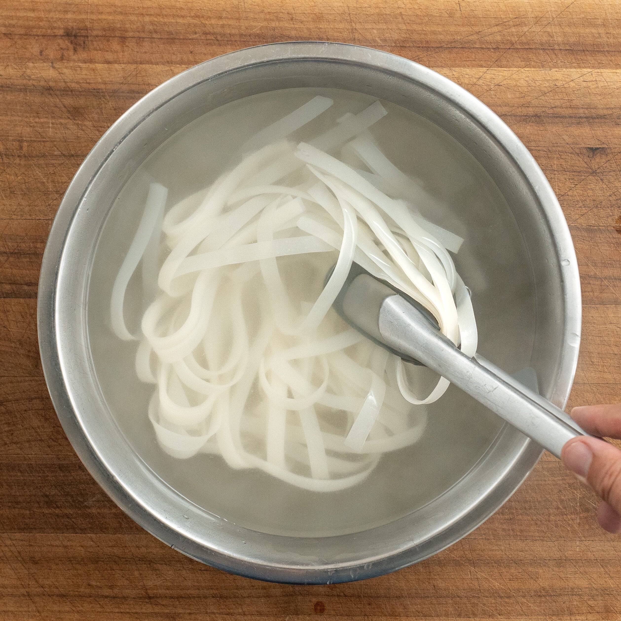 stirring wide rice noodles in silver bowl with tongs