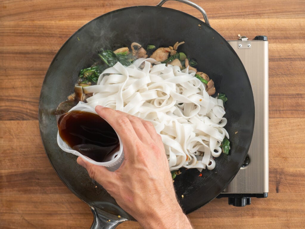 drizzling brown liquid on top of rice noodles and chicken in wok