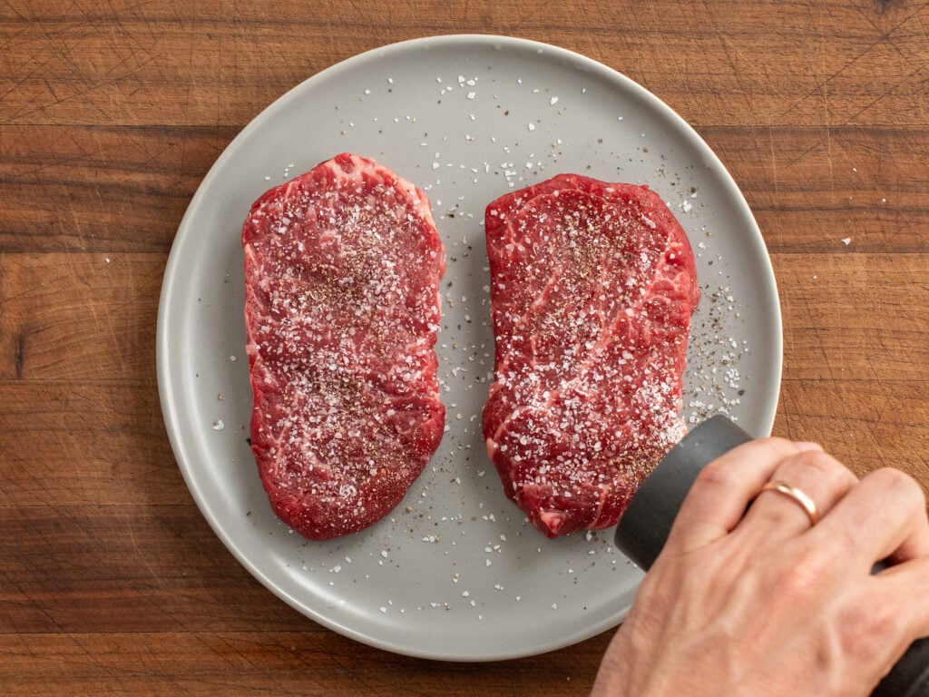 pepper grinder adding pepper to two pieces of raw steak on grey plate