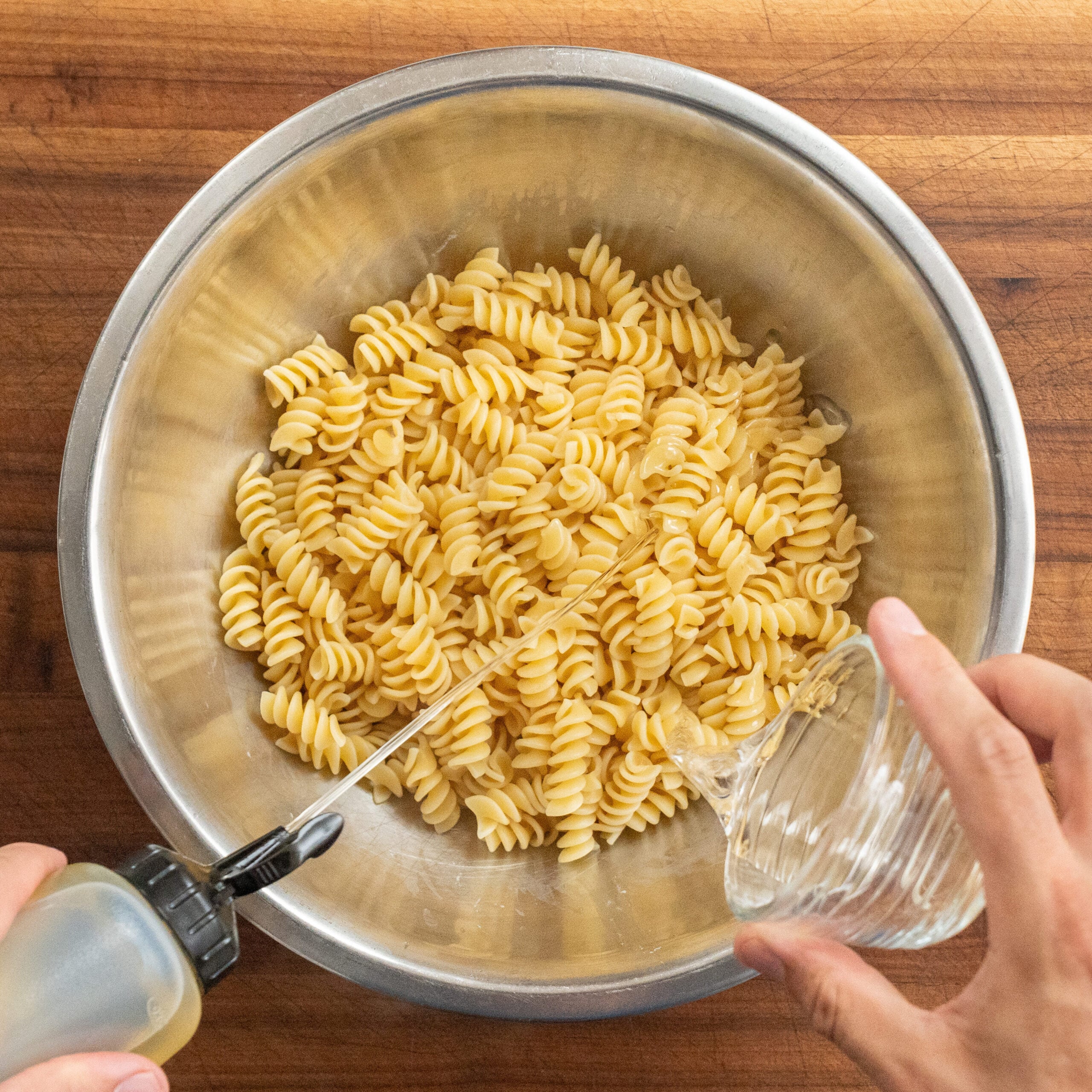 adding oil and vinegar to cooked fusilli pasta in silver bowl