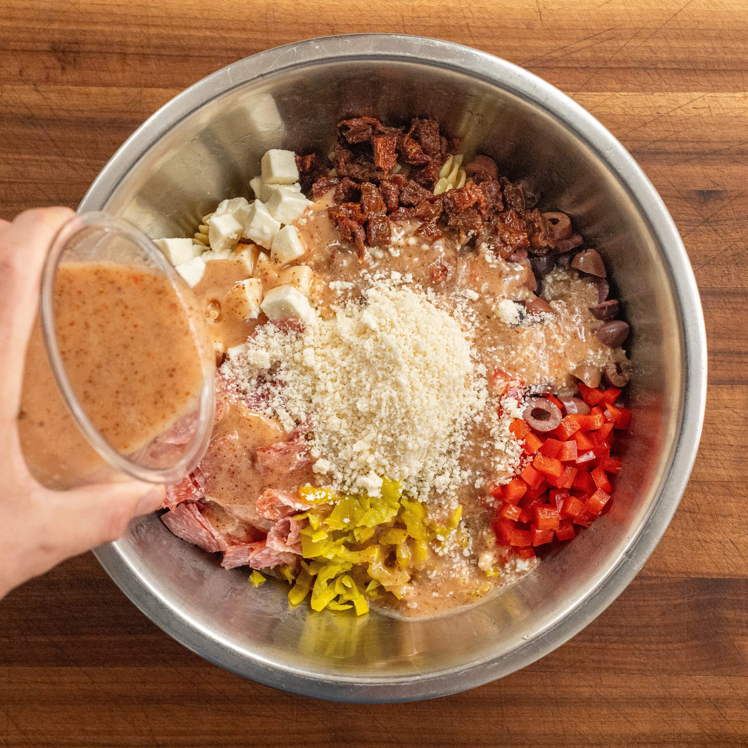 pouring Italian dressing over pasta salad ingredients in metal bowl