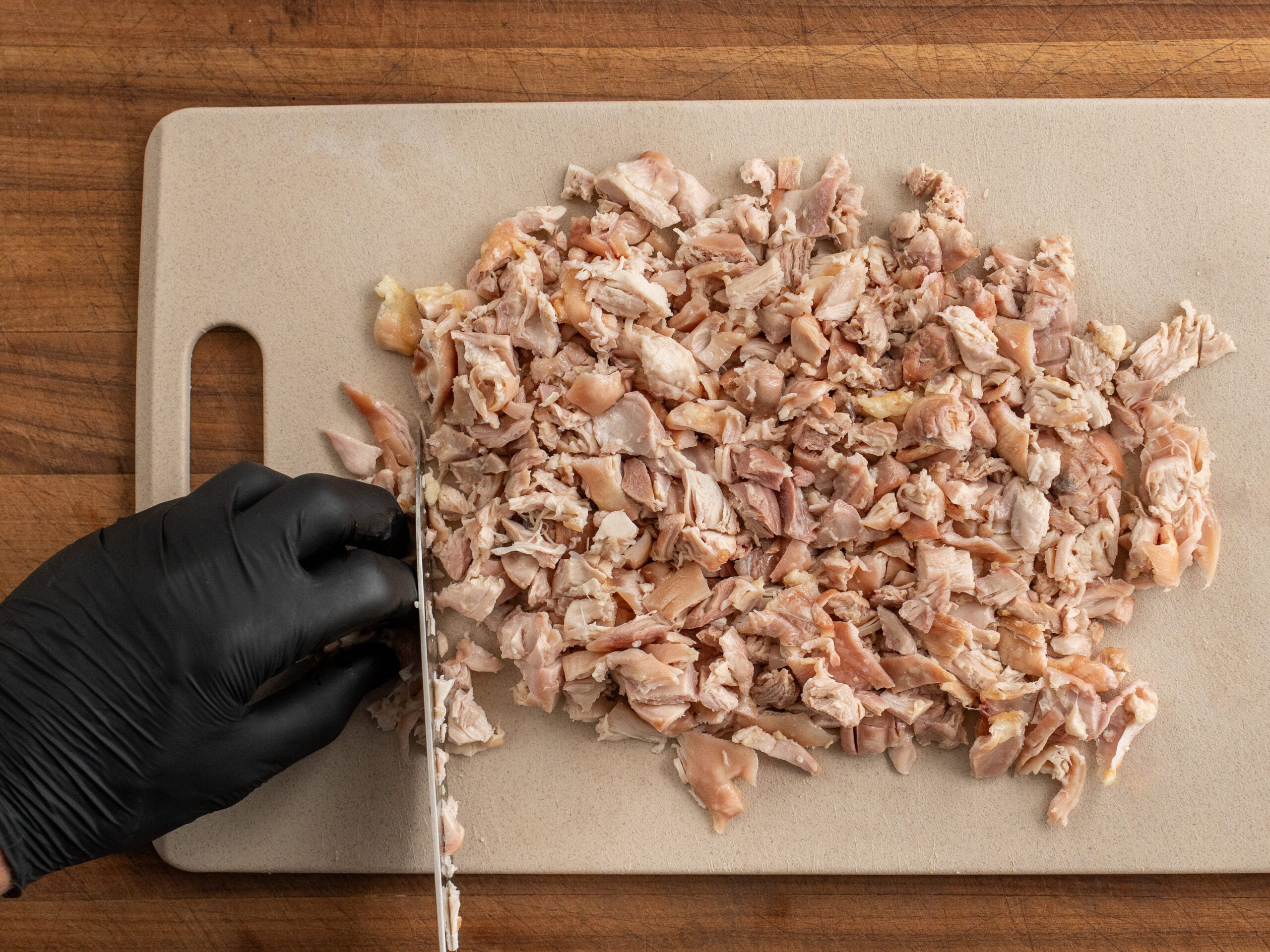 black gloved hand chopping chicken thighs on brown cutting board