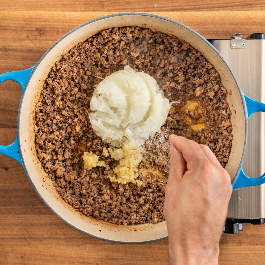 hand adds pinch of salt to grated onion and minced garlic sitting on top of cooked ground beef in blue dutch oven