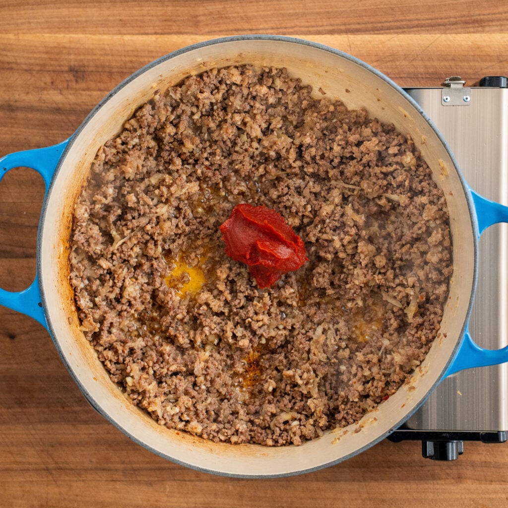 spoonful of tomato paste sitting on top of cooked ground beef in blue dutch oven