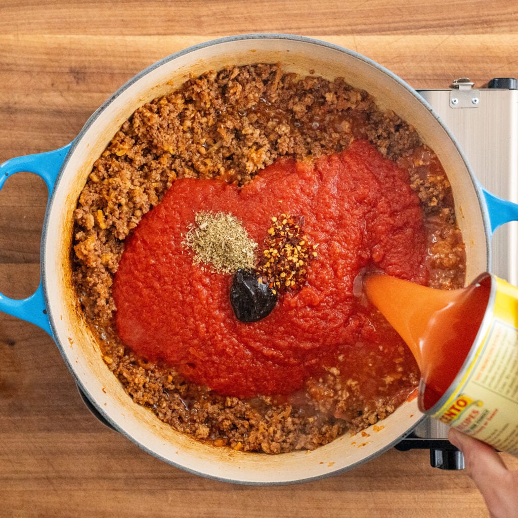 adding can of water to crushed tomatoes bouillon paste dry herbs sitting on cooked ground beef in blue dutch oven