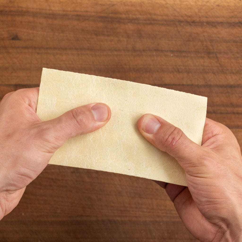 hands holding dry sheet of lasagna over wood cutting board