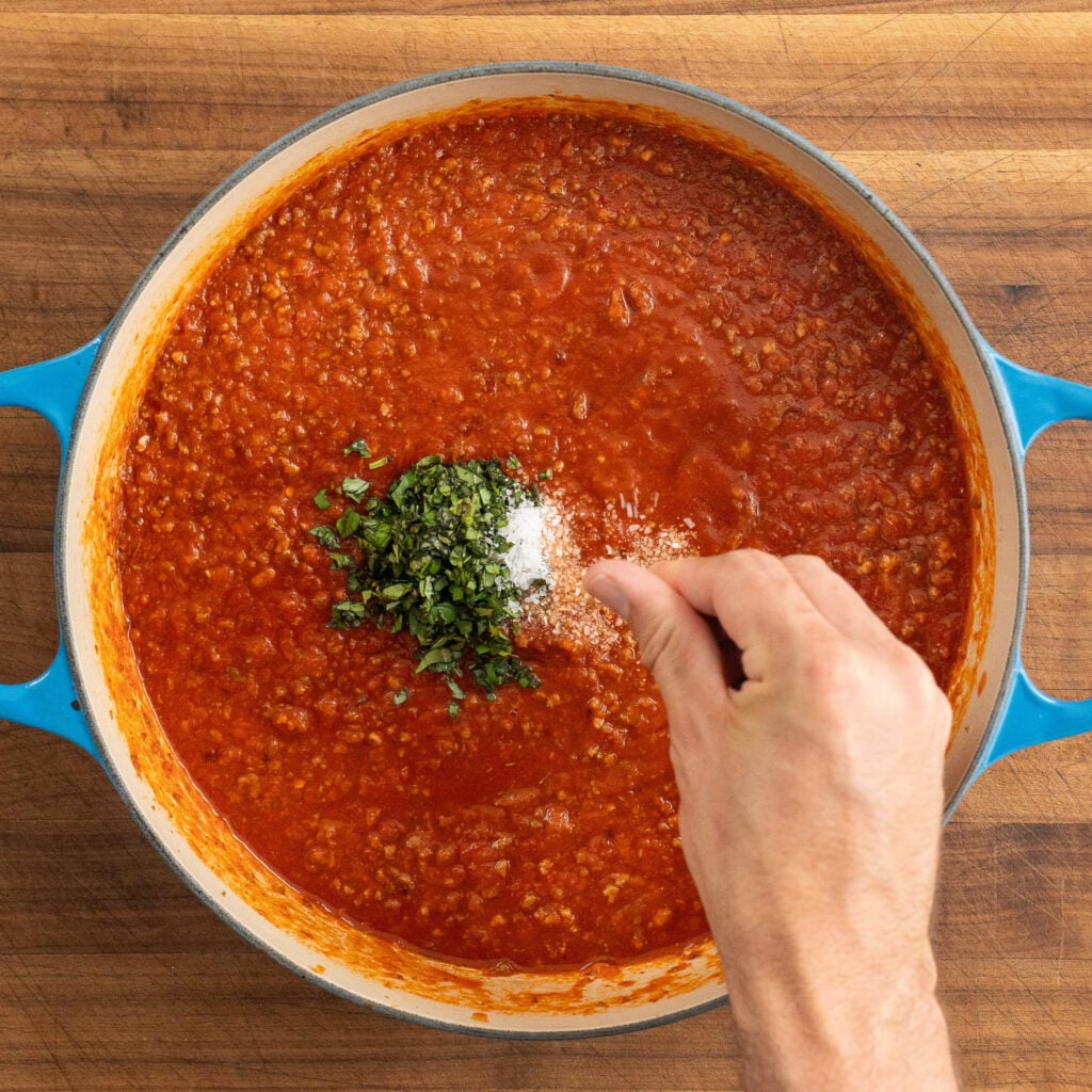 hand sprinkles salt into simmering tomato meat sauce