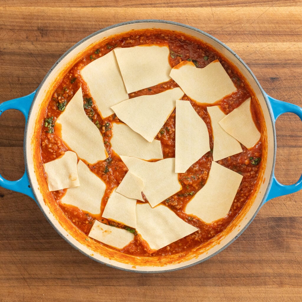 broken pieces of lasagna sheet arranged to cover top of meat sauce in blue dutch oven