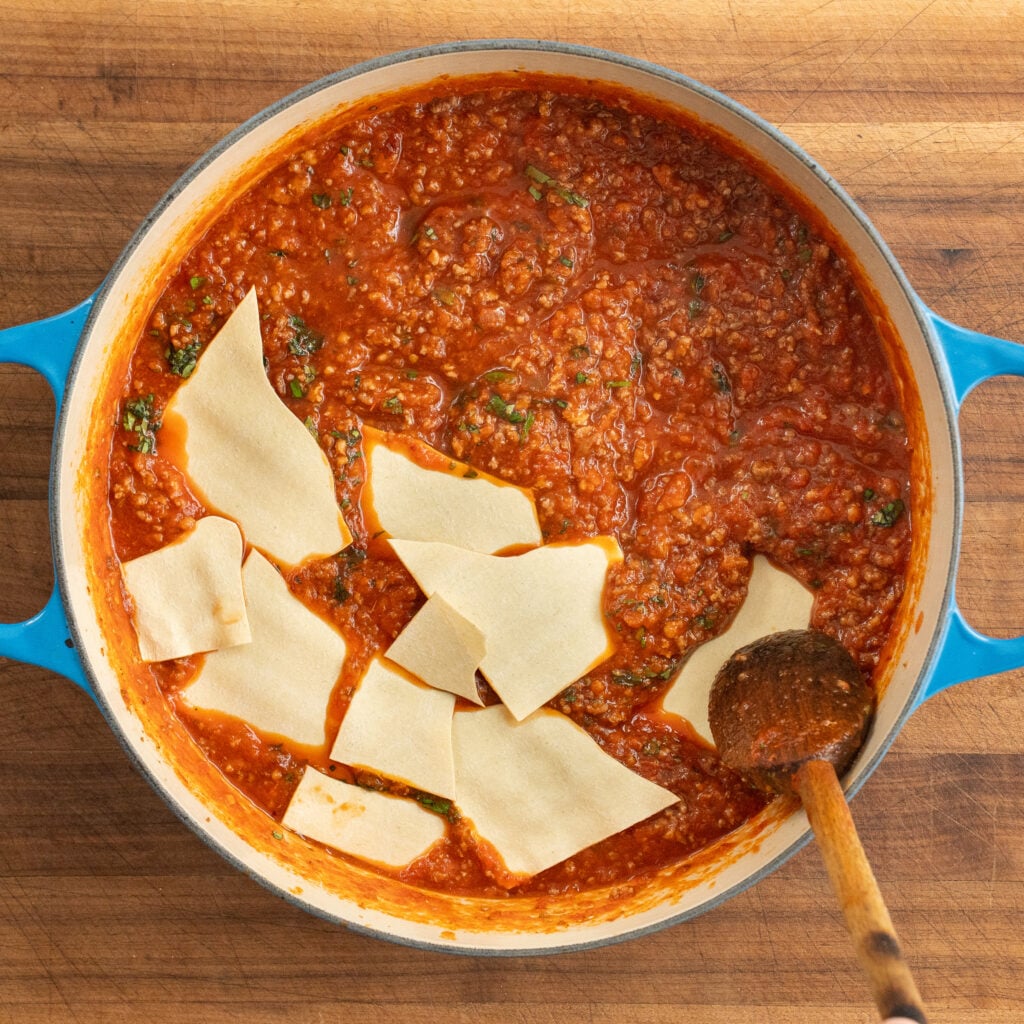 wooden spoon pressing to submerge broken lasagna sheet pieces into meat sauce