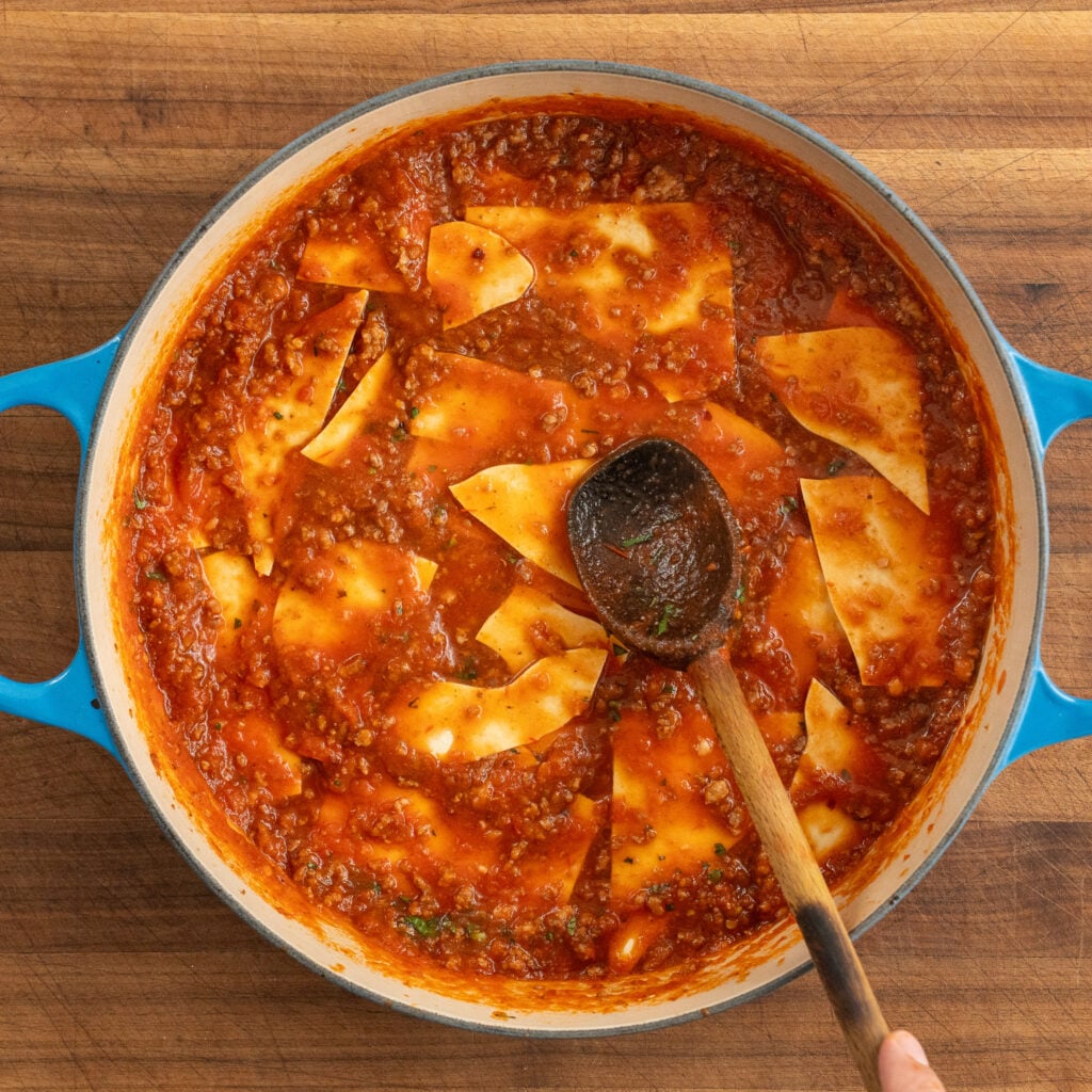 wooden spoon covering pieces of lasagna sheet with meat sauce
