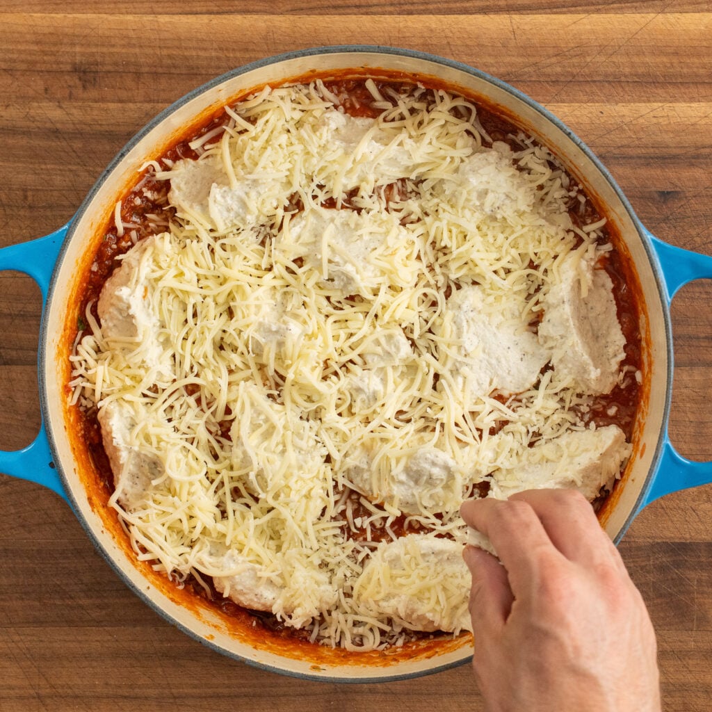 hand sprinkles shredded mozzarella on blue dutch oven filled with ricotta and meat sauce