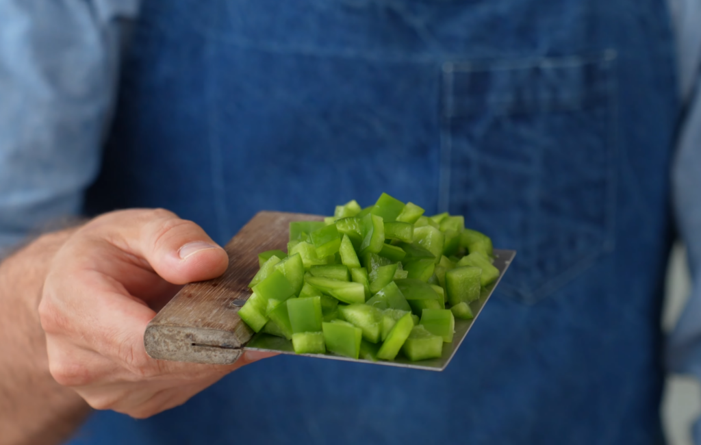 man in blue apron holding metal bench scraper with diced green peppers on it
