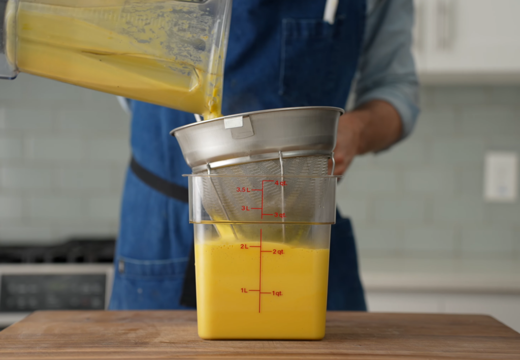 man in blue apron pouring yellow soup through fine mesh strainer into 4qt clear container