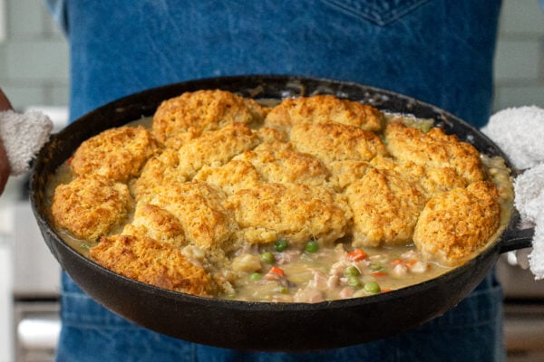 man in blue apron holding cast iron pan full of drop biscuit chicken pot pie